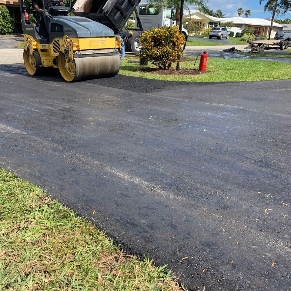 Road roller compacting fresh asphalt on a driveway, with a dump truck, grass, and houses in the background.