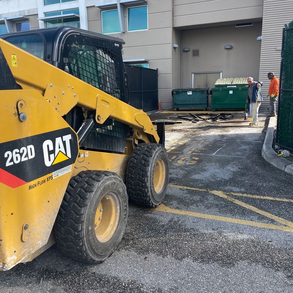 Yellow CAT 262D skid steer near green dumpsters with two workers outside a building.