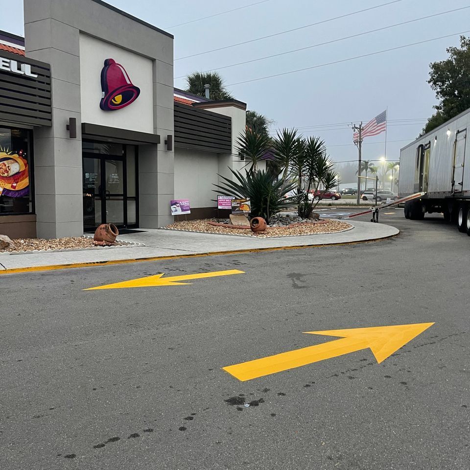 Taco Bell restaurant exterior with yellow directional arrows on pavement and a semi-truck parked in the background.