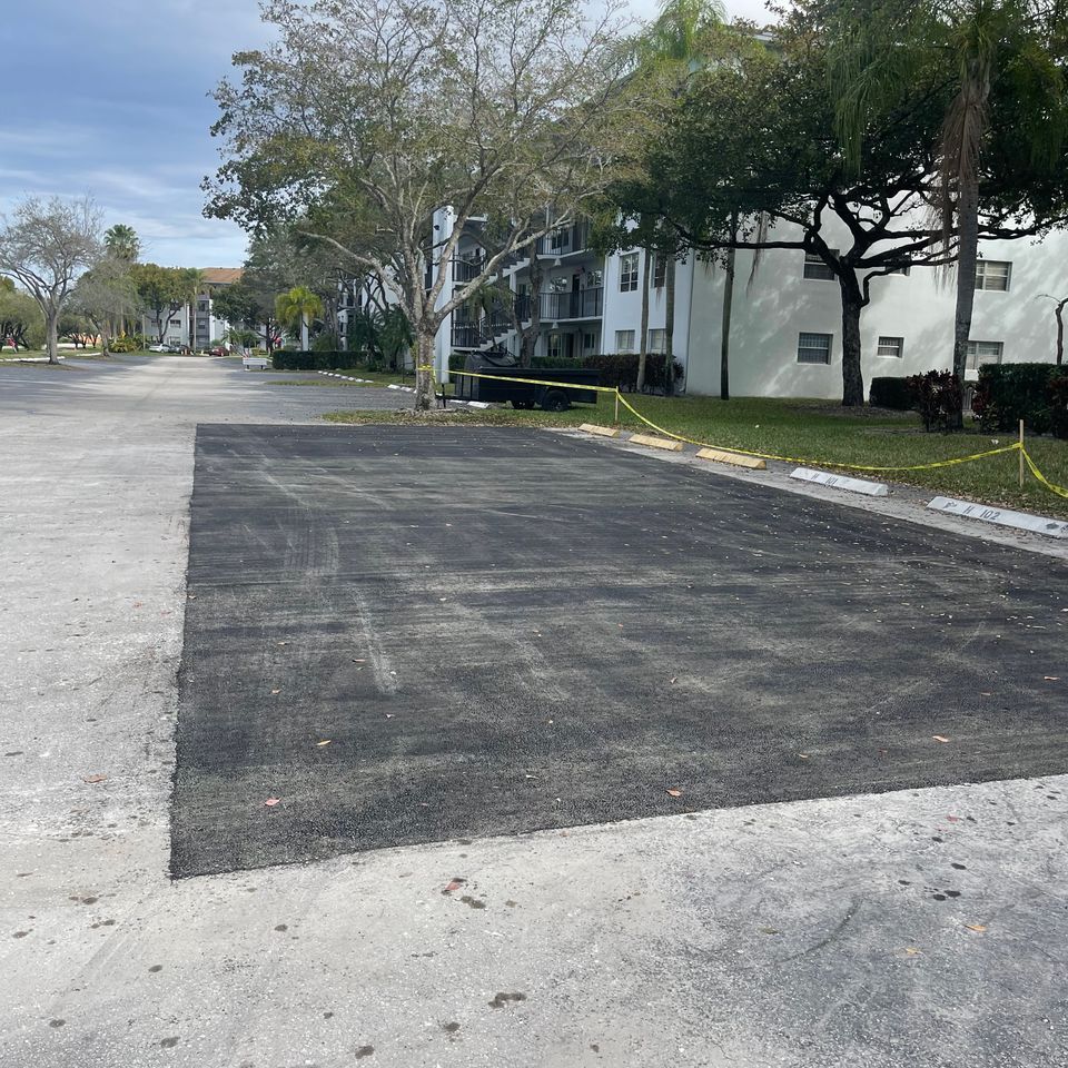Newly paved asphalt parking area next to existing gray pavement, with buildings and trees in the background.