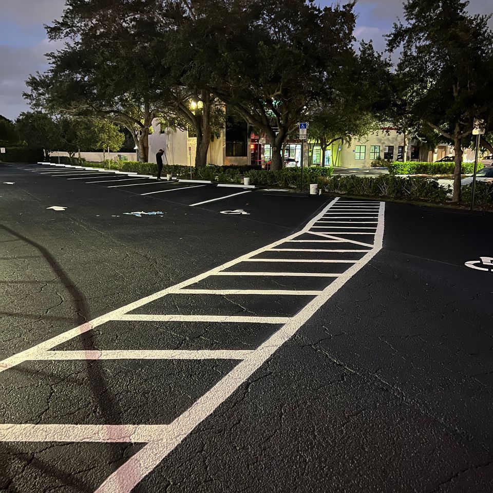 Asphalt parking lot at night with painted white lines, including a crosswalk and parking space markings. Buildings in the background.