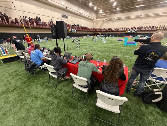 A group of people are sitting at tables in an indoor stadium watching a drone race.