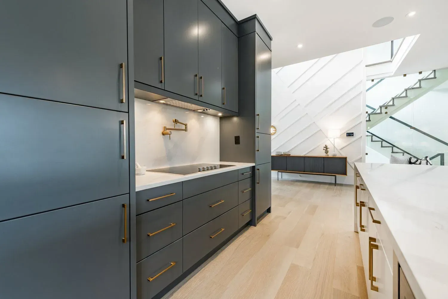 A kitchen with gray cabinets and white counter tops and a staircase in the background.