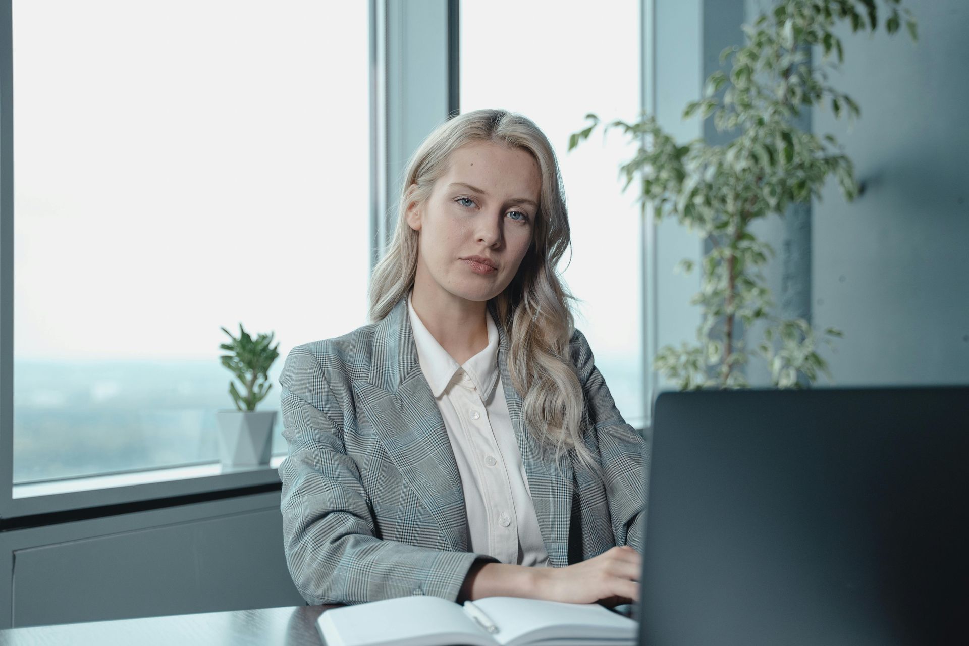 Blonde woman in blazer at desk, looking at camera. Laptop, notebook, and plant visible. Windows in background.