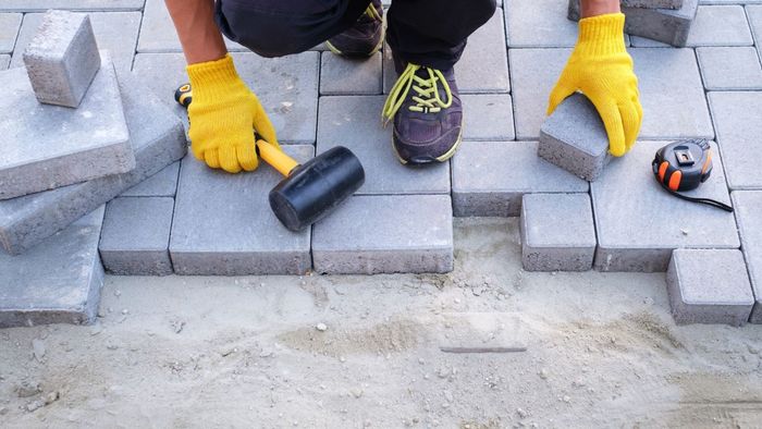 Blocks being installed for a block paving driveway. The blocks are grey and the man installing has yellow gloves on his hands.