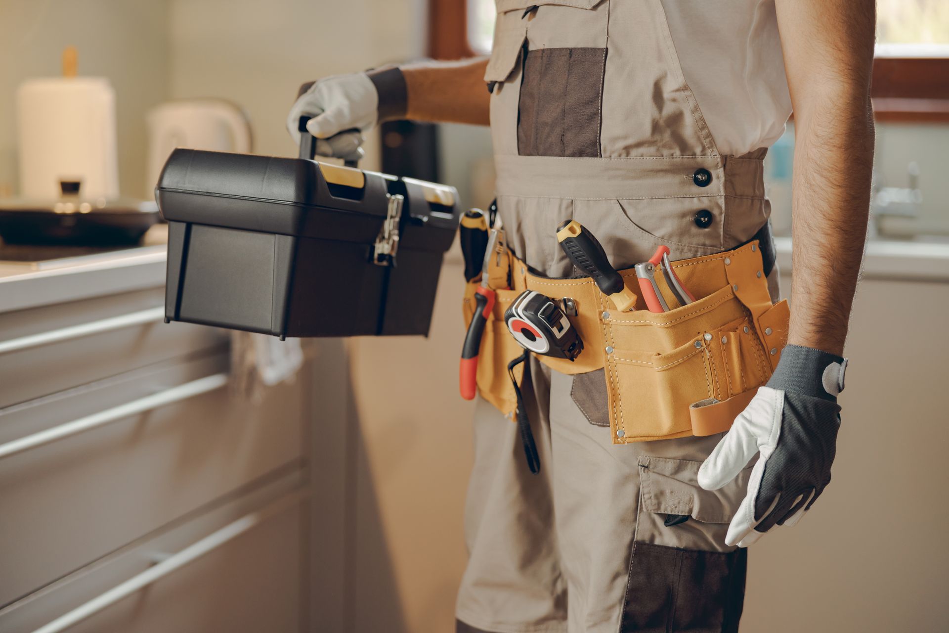 A worker in beige overalls and gloves holds a black toolbox in a kitchen, wearing a tool belt with various instruments.