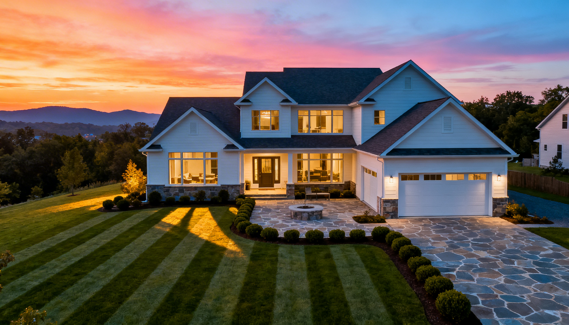 A two-story white house with a stone patio and striped lawn sits against a vibrant, multi-colored sunset sky.