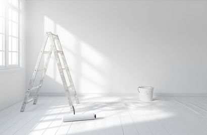 A room with white walls, a step ladder, a paint roller on the floor, and a paint bucket, bathed in sunlight.