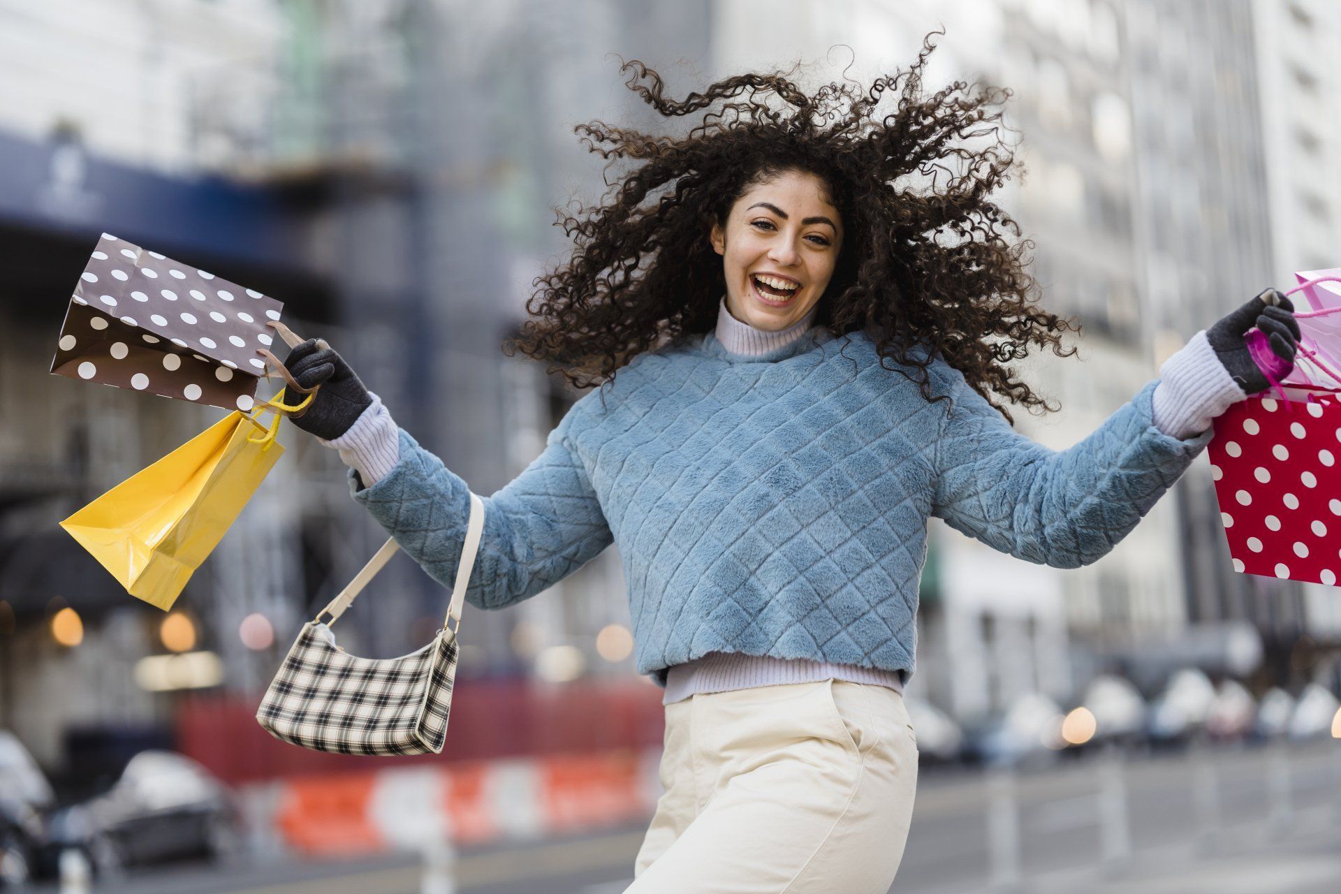 Woman with curly hair, arms outstretched, holding shopping bags on a city street; joyful expression.