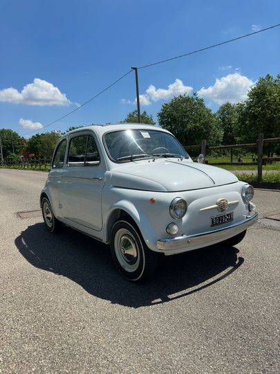 Una piccola auto d'epoca bianca parcheggiata su una strada sterrata sotto un cielo azzurro con alberi sullo sfondo.