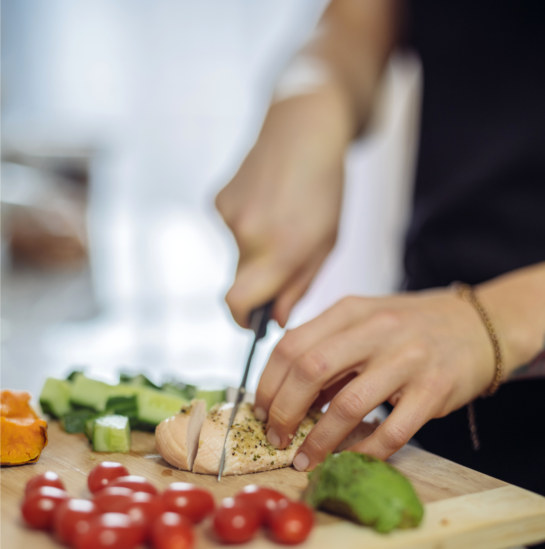 Person slicing chicken breast on a wooden cutting board with vegetables.
