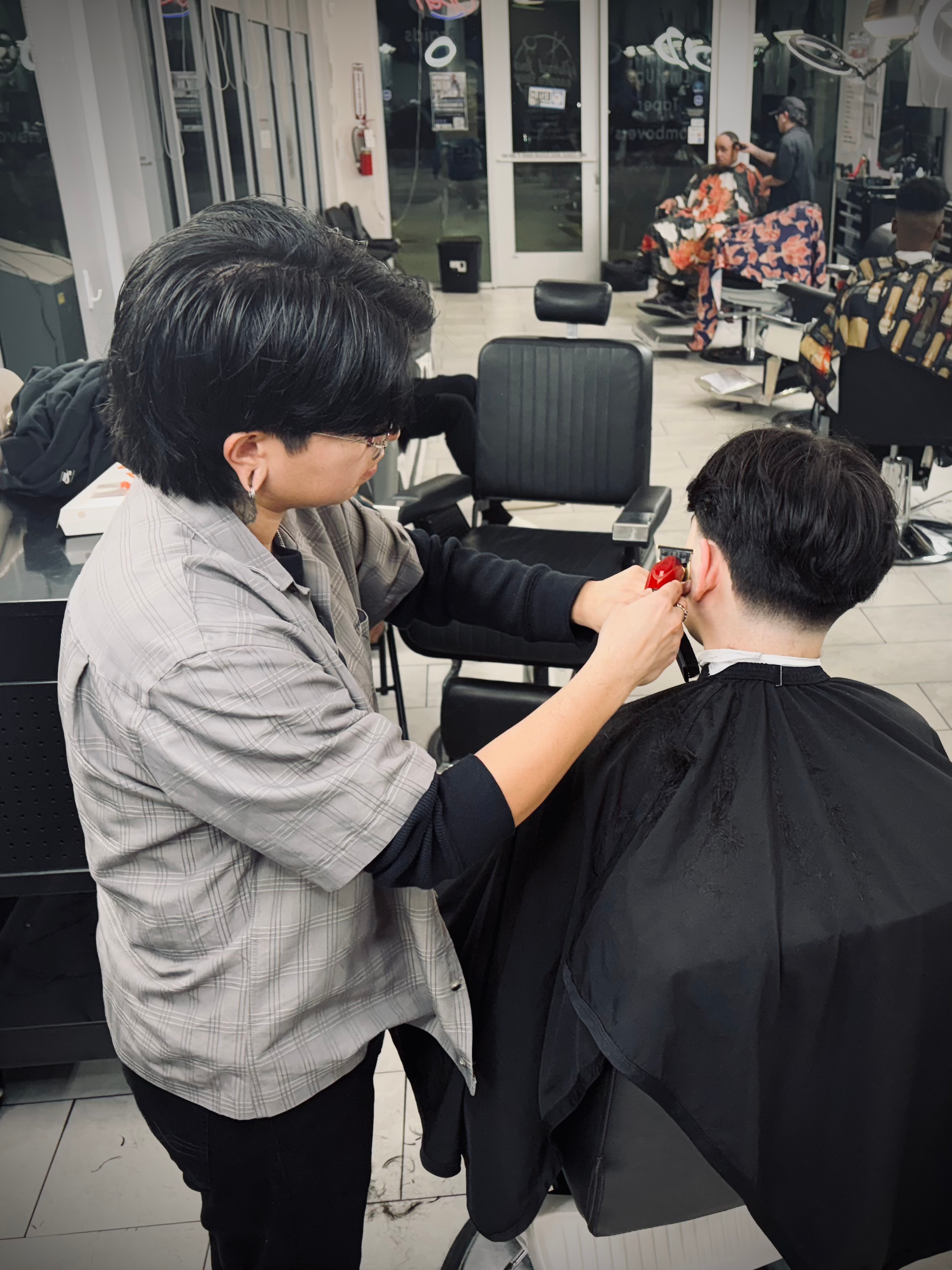 A barber in a gray shirt uses electric clippers to trim the hair of a seated client wearing a black cape in a salon.
