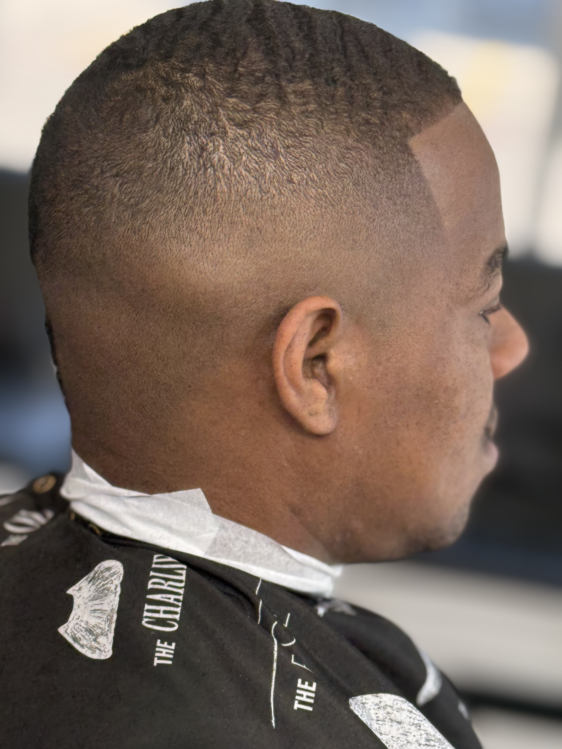A profile view of a man with a short, precision-faded haircut, wearing a barber’s cape.