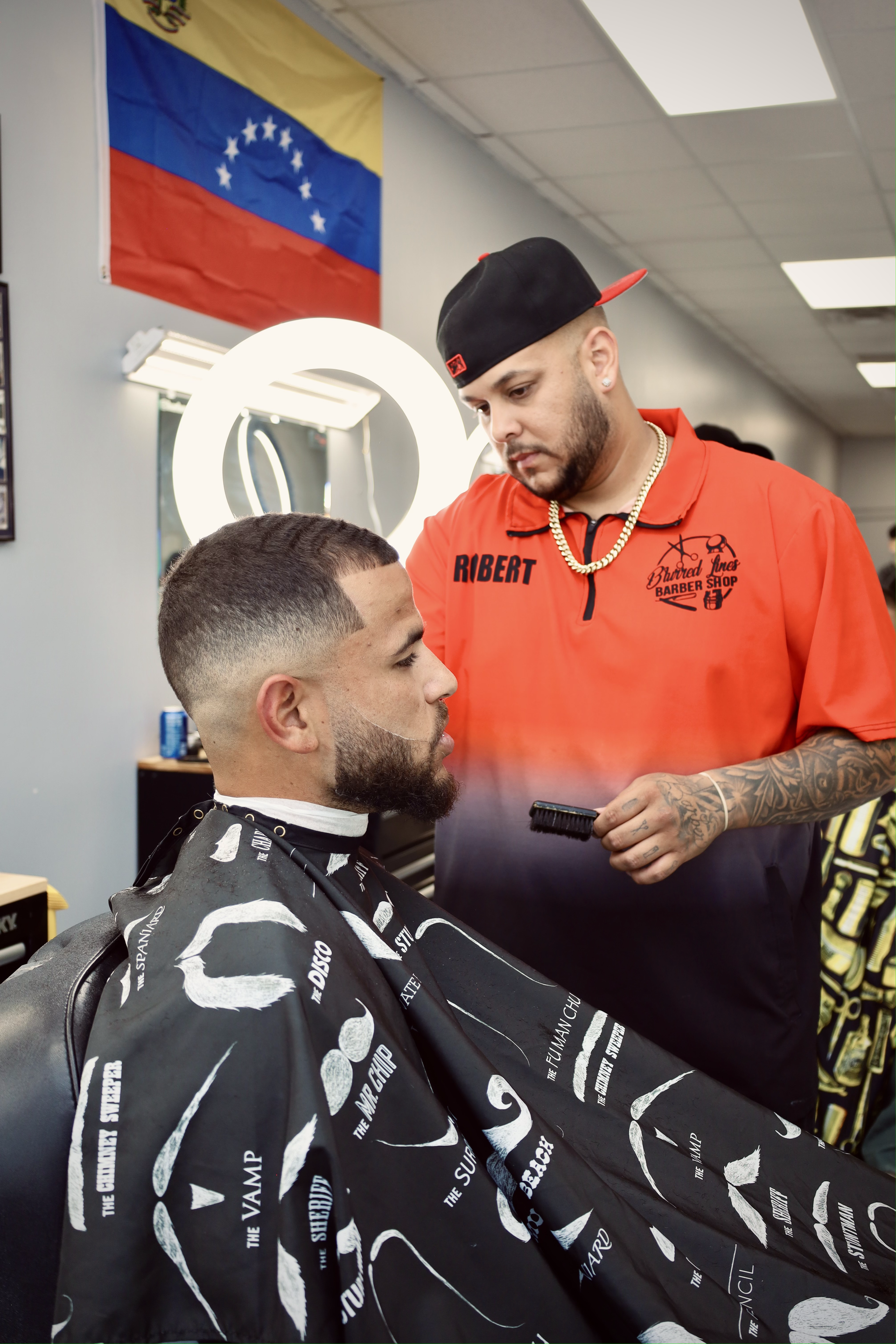 A barber in an orange shirt cuts a client’s hair in a shop with a Venezuelan flag displayed on the wall.