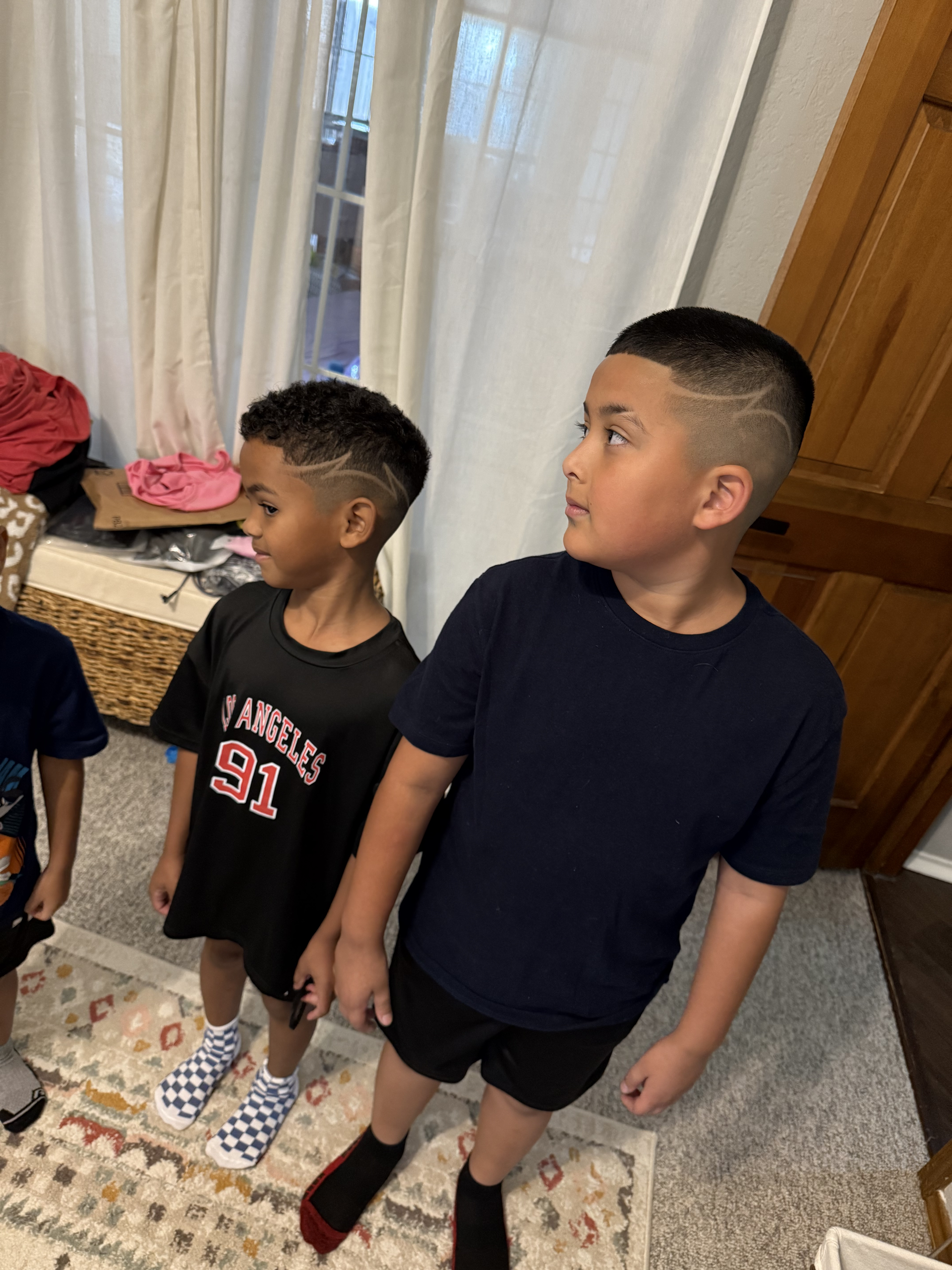 Two children standing indoors, wearing shorts and shirts, looking to their left with styled hair featuring shaved designs.