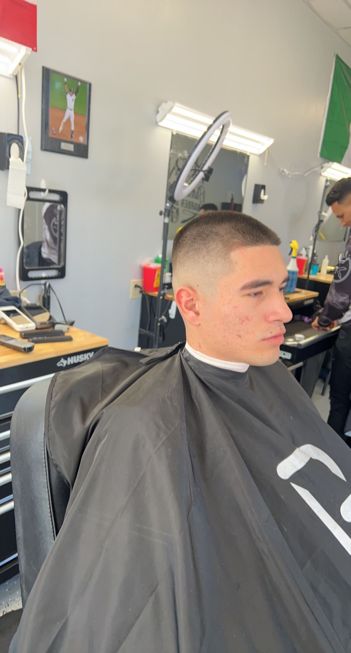 A person sits in a barber chair wearing a black cape after receiving a fresh buzz cut with a skin fade.