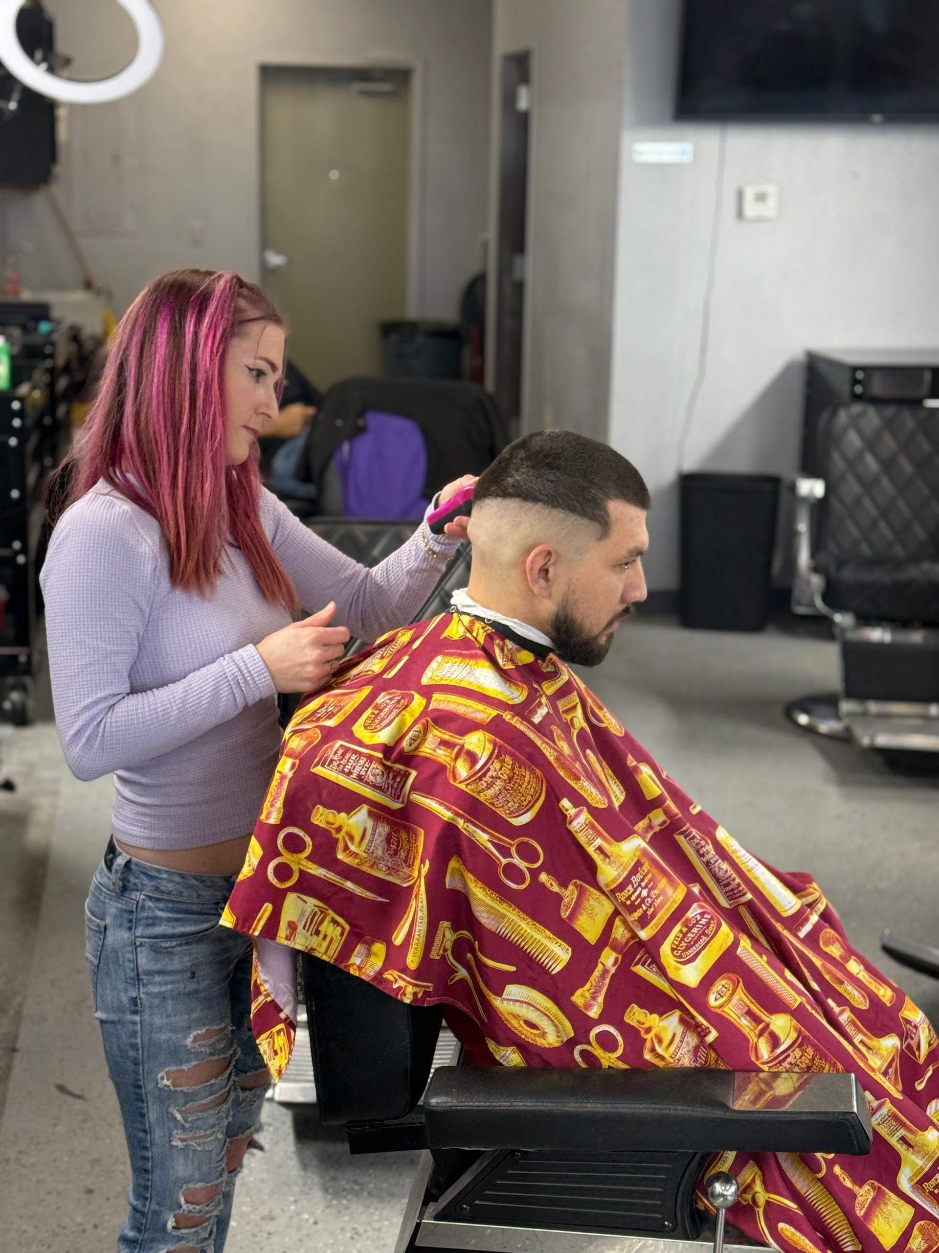 A stylist with pink hair gives a person a haircut in a salon, using clippers on the side of their head.