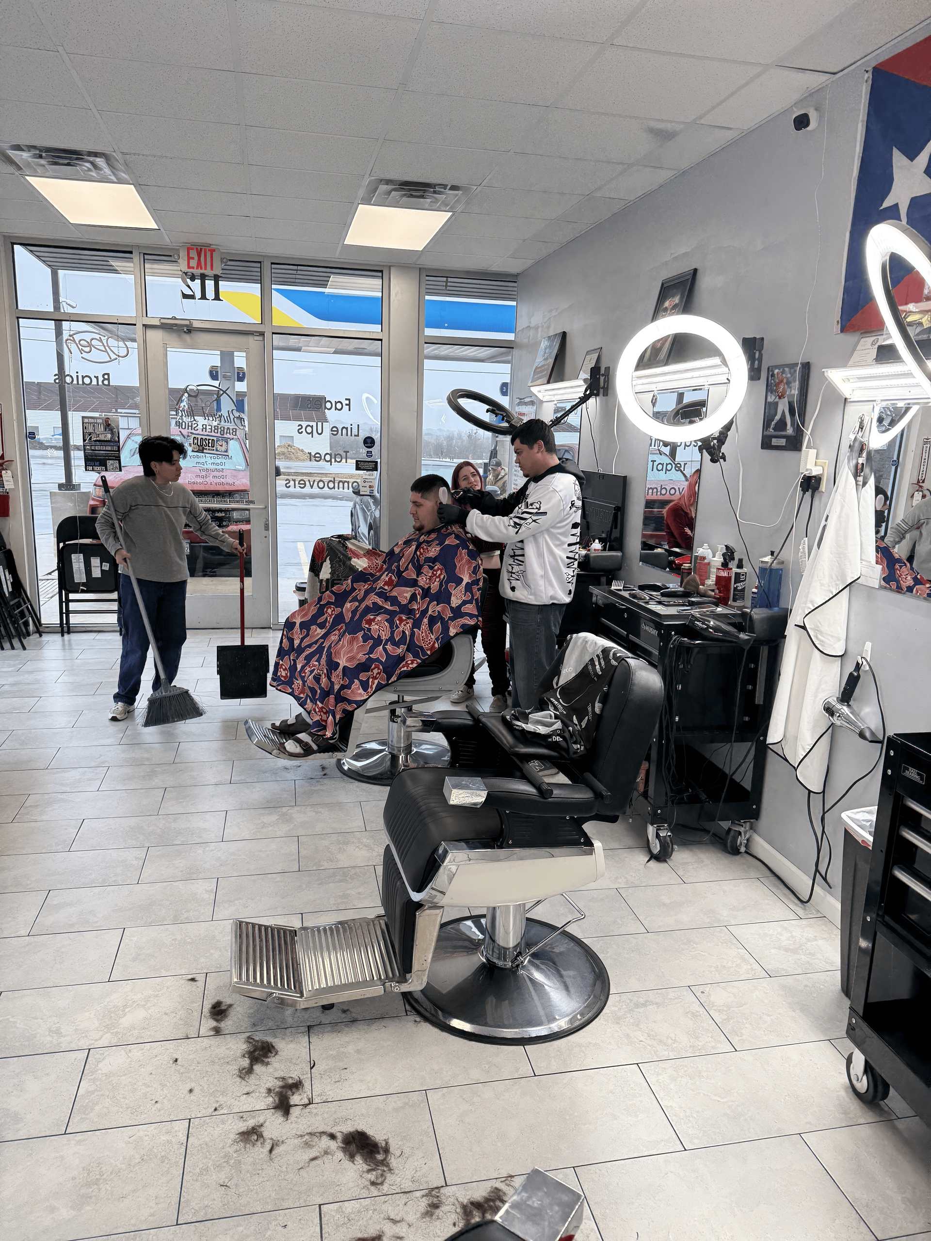 A barber cuts a client’s hair in a brightly lit shop, while another person sweeps hair from the tiled floor nearby.