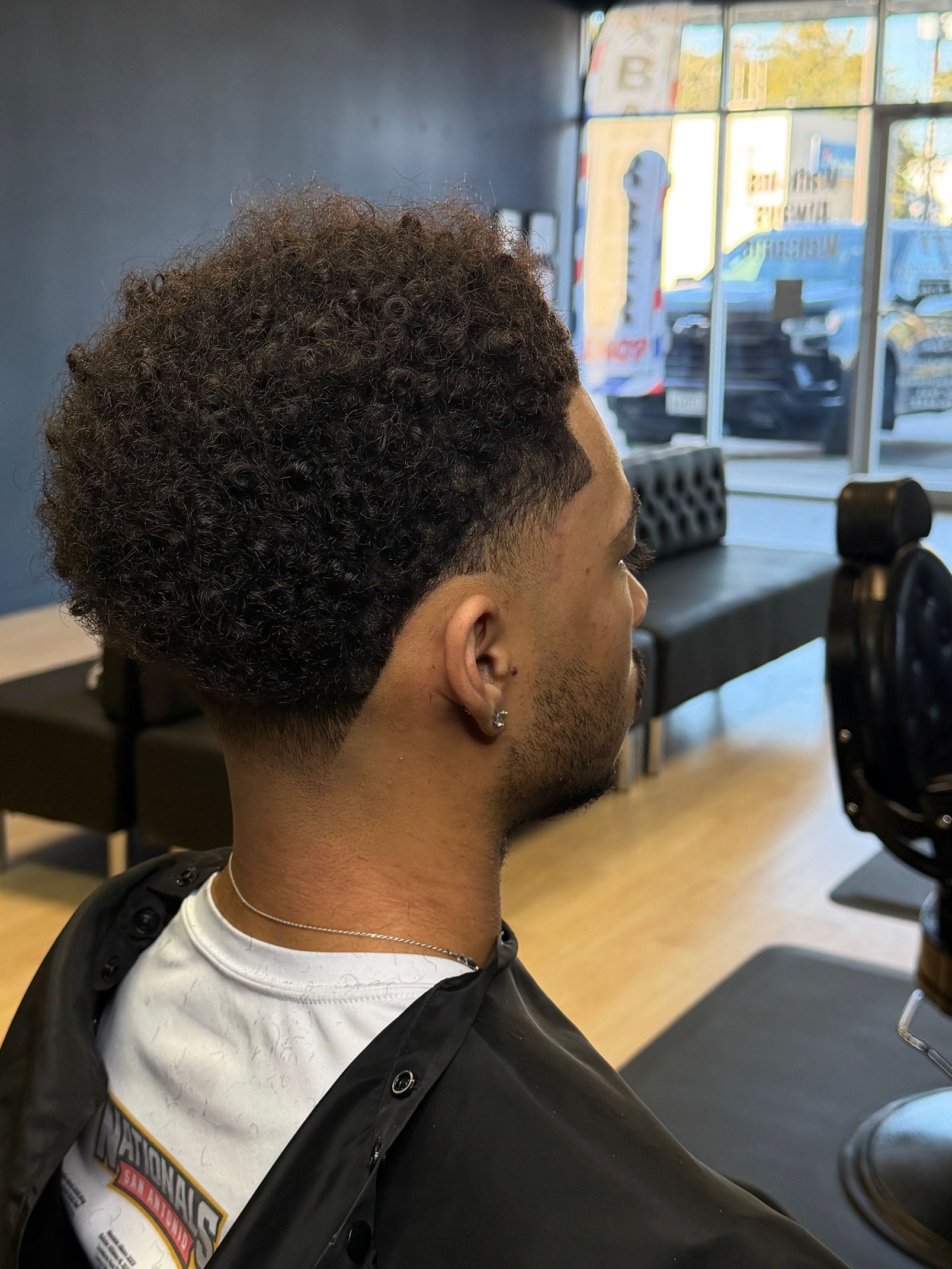 A side view of a person with a short, curly-textured haircut with a faded neckline, sitting in a barbershop chair.