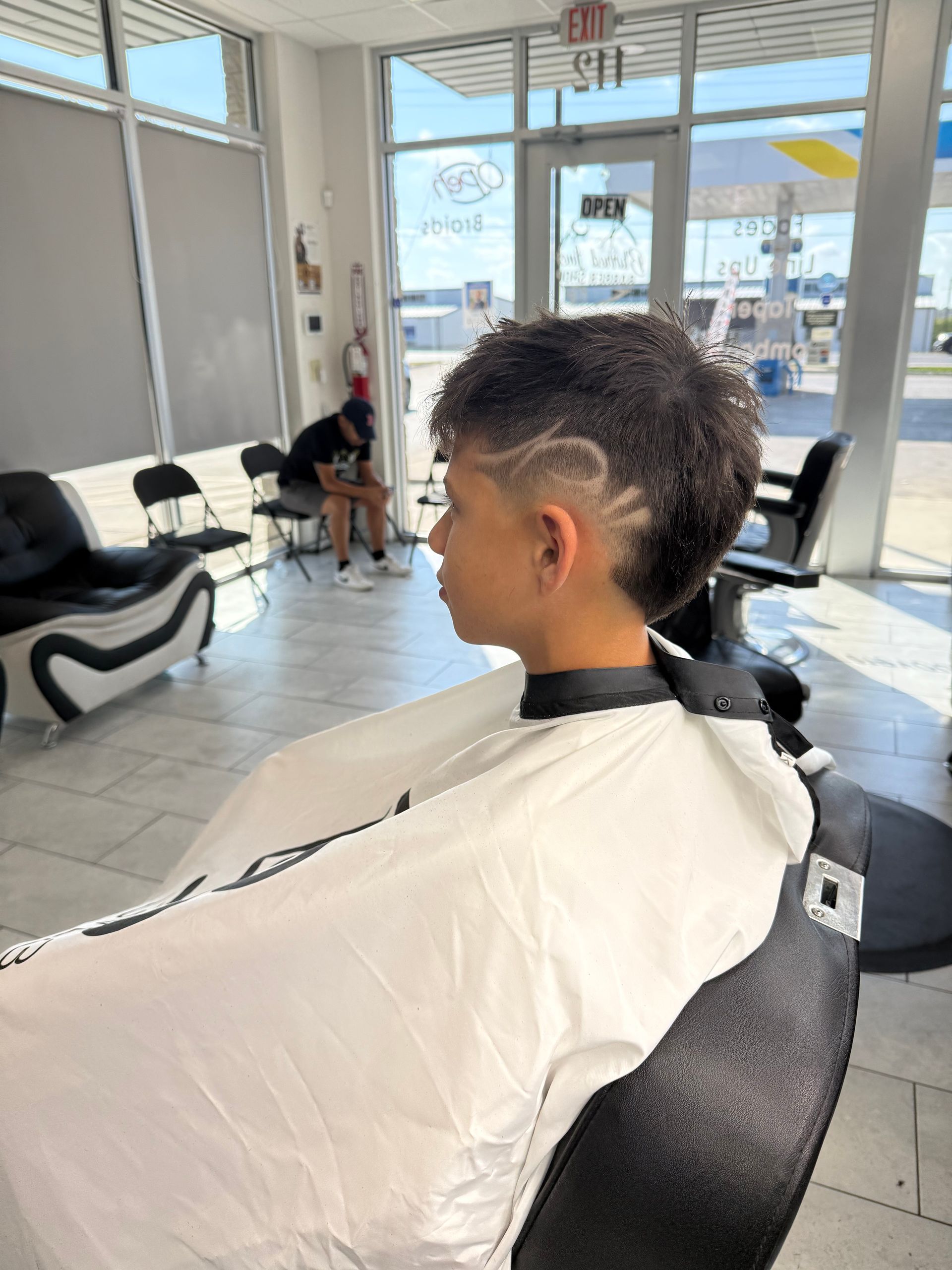A person sits in a salon chair wearing a white cape, displaying a short, textured haircut with a shaved design on the side.