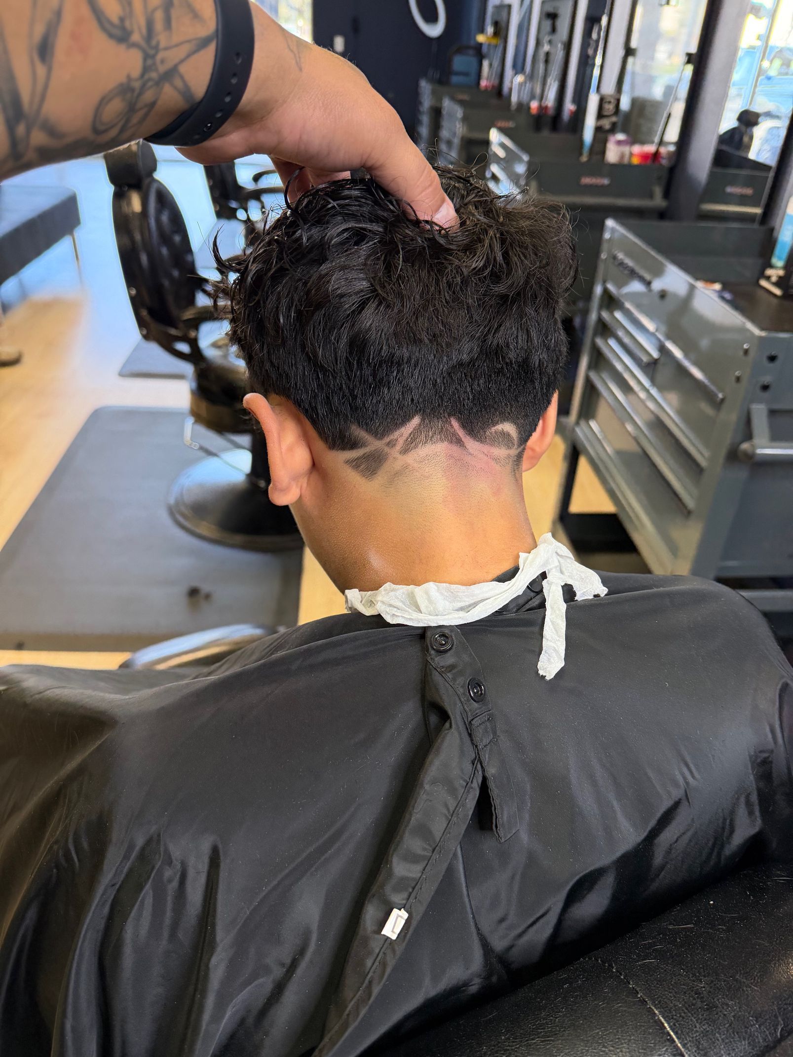 A barber’s hand rests on the back of a client's curly-haired head, showing a fresh haircut with a faded neckline.
