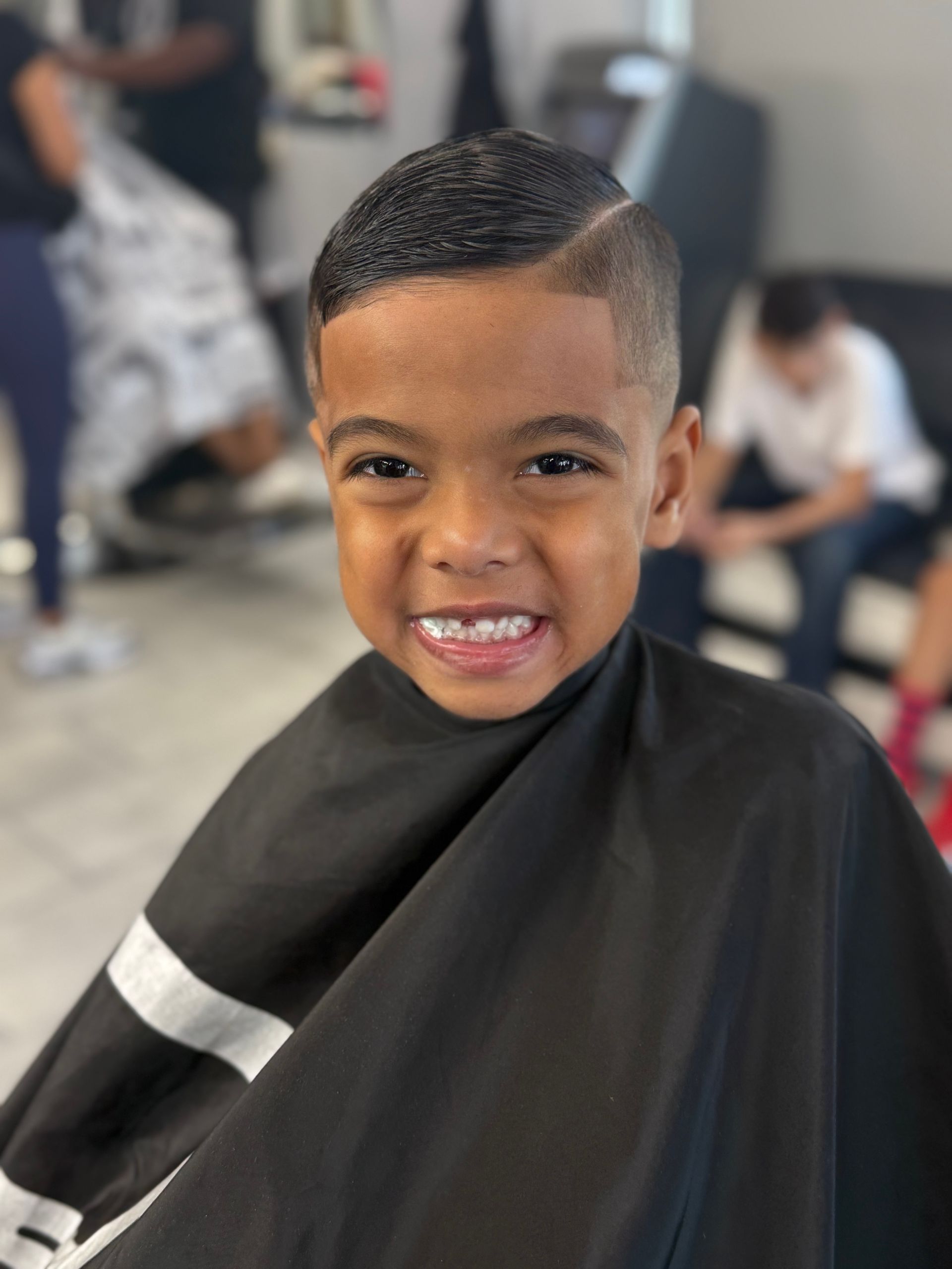 A smiling child in a black salon cape, showcasing a fresh haircut with a clean-shaven side part in a barbershop.