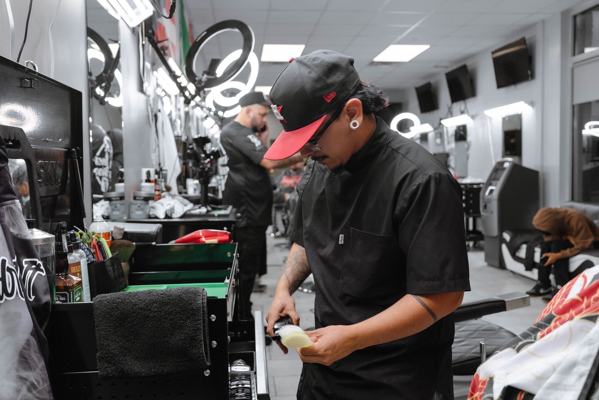 A barber in a black uniform prepares a tool at a station in a brightly lit barbershop with multiple chairs and lights.