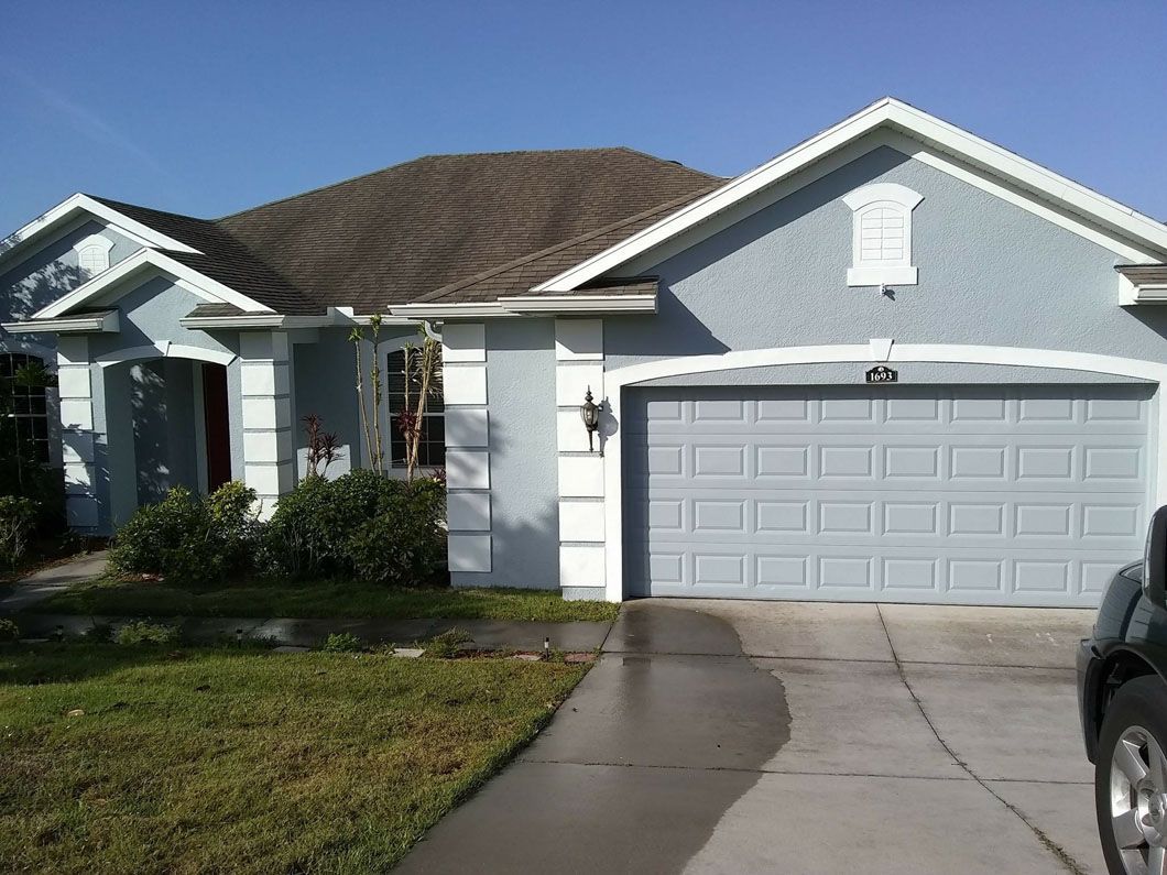 Blue stucco house with gray garage door and driveway, green lawn, and brown roof
