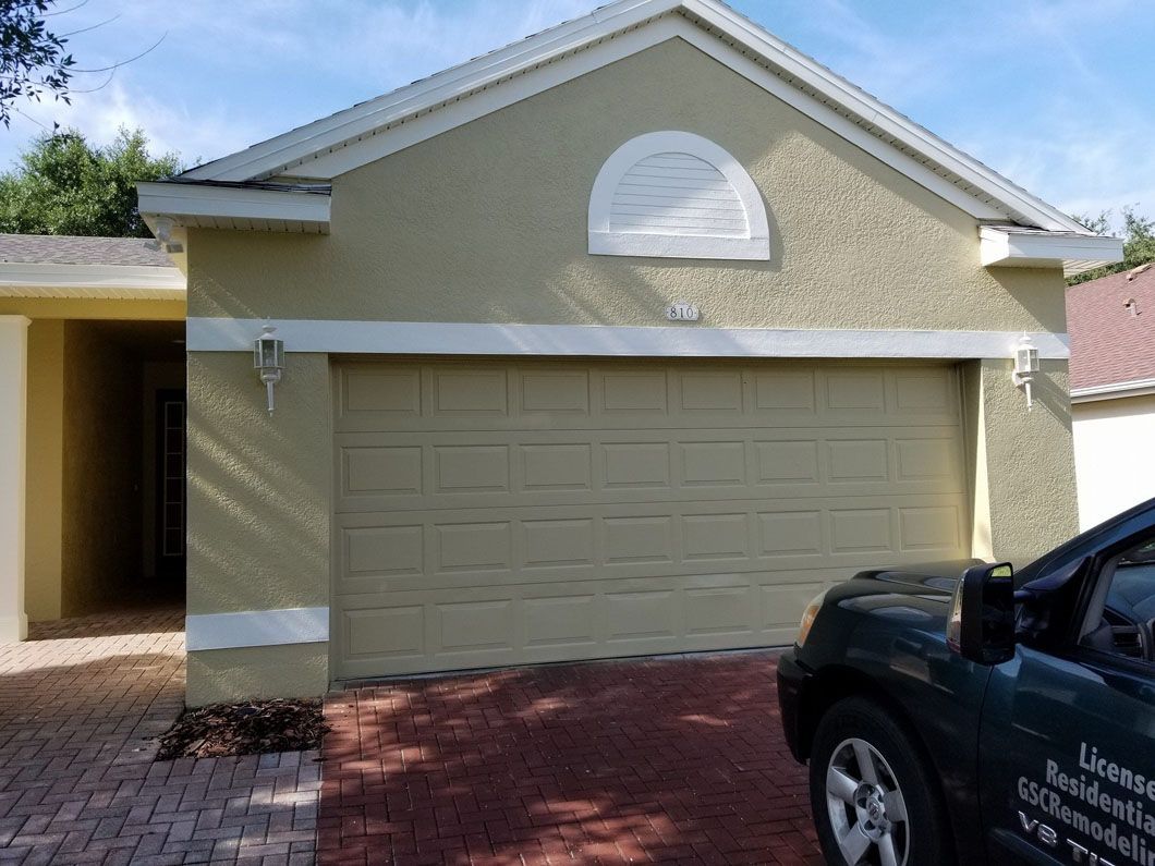 Tan stucco house with garage, brown brick driveway, and dark SUV in the right corner