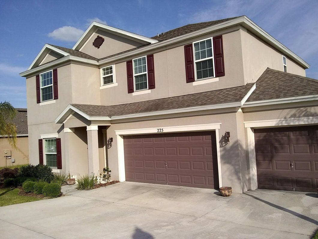 Two-story beige house with brown shutters, garage doors, and roof under a blue sky