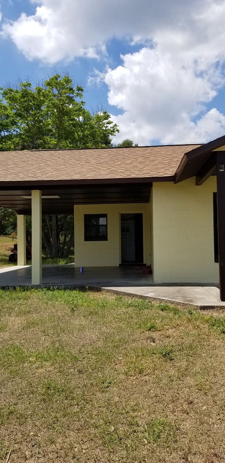 A light yellow building with a covered porch, brown roof, and gren grass, under a blue sky.