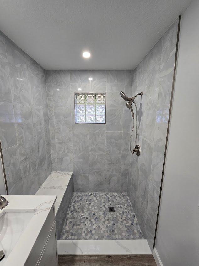 Accessible walk-in shower with gray tiled walls and floor, showerhead, bench, and small window