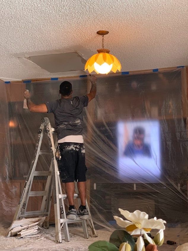 Man on a ladder paints a wall with plastic sheeting. Yellow ceiling light, white ceiling