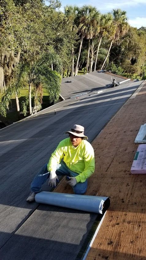 Roofer on a roof, rolls out roofing material. Wearing a safety hat and bright long-sleeve shirt. Sunny day, palm trees.