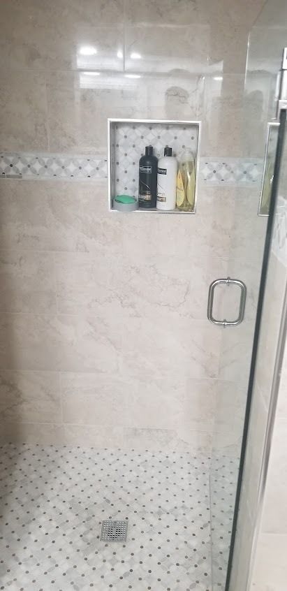 Shower with light-colored stone tiles, mosaic floor, glass door, and built-in shampoo niche