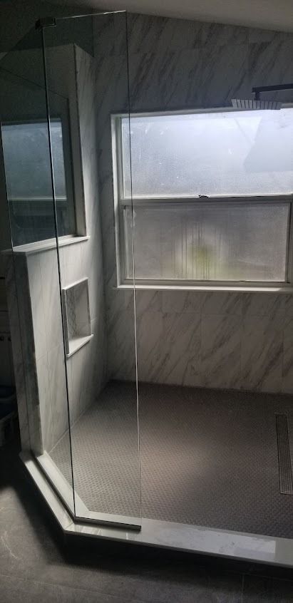 A shower with a clear glass door, gray tiled walls, and a window