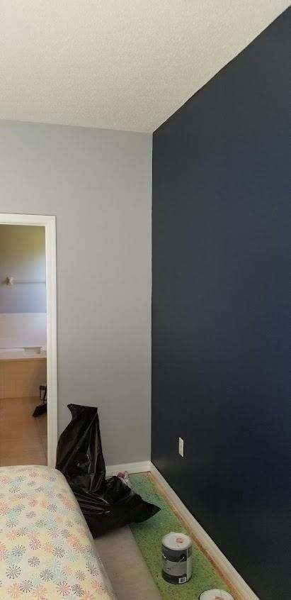 A bedroom corner with walls painted gray and dark blue, a bed, and painting supplies