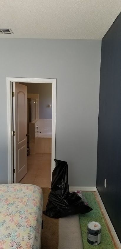 Bedroom with gray and dark blue walls, open door to bathroom, trash bag, paint can, bed