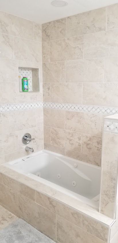 A beige-tiled bathroom with a built-in tub. There is a recessed shelf with a bottle.