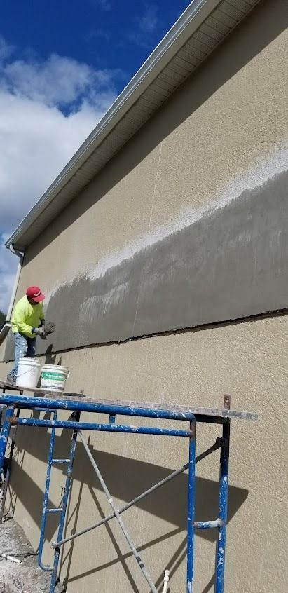 A worker applies stucco to a building's exterior. He stands on a scaffold, wearing a hard hat.