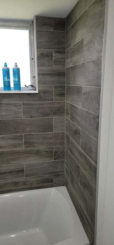 Bathroom with gray wood-look tile. A white tub is below a window with two blue bottles on the sill