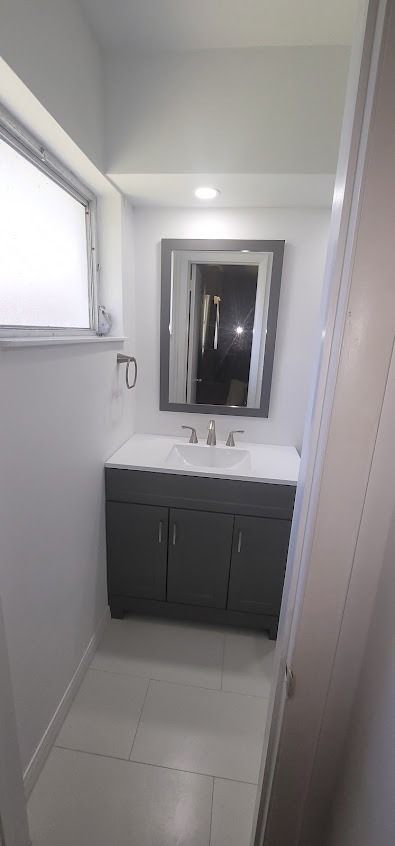 Bathroom with gray vanity, mirror, and white walls. Light from a window.
