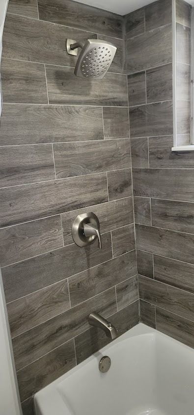 Bathroom shower with gray wood-look tile, stainless steel fixtures, and a white tub