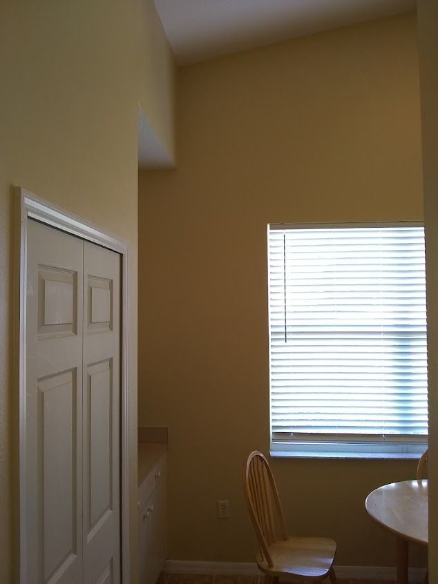 Interior view of a room with a closet, window, chair, and table, with tan walls and a high ceiling