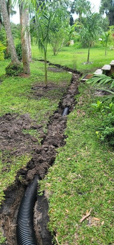 A trench dug in grass contains a black drain pipe, with trees and green vegetation in the background