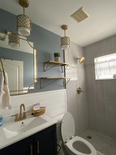 Bathroom with blue wall, white tile, gold fixtures, vanity, toilet, shelves, and pendant lights
