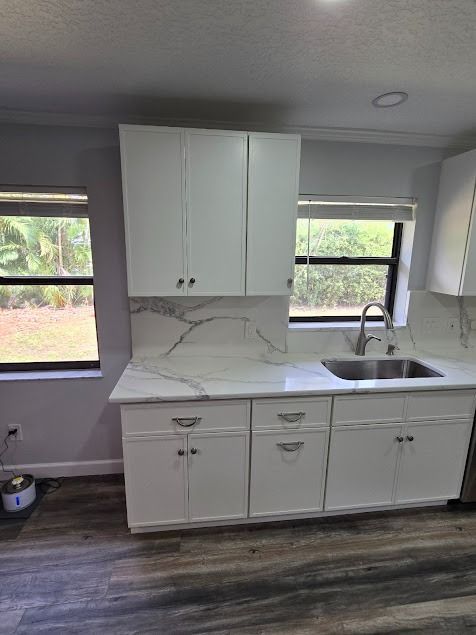 White kitchen cabinets with marble countertops and a stainless steel sink, next to windows and dark floors
