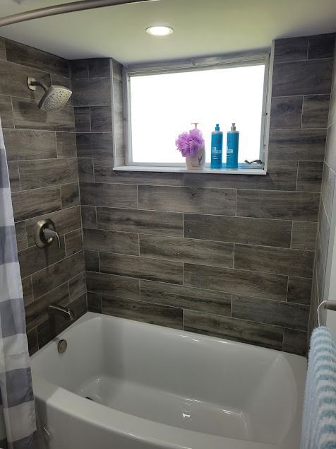 Bathroom with brown tile, white tub, and a window with toiletries