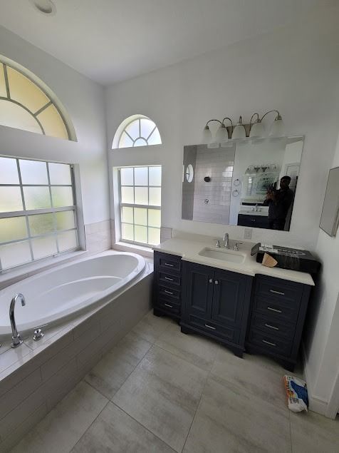 Bathroom with tub and vanity, featuring navy blue cabinets, white counter and tile. Includes two arched windows