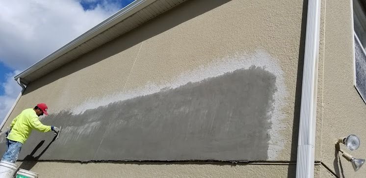 Man applying stucco to a wall outdoors, visible clouds, and part of the house